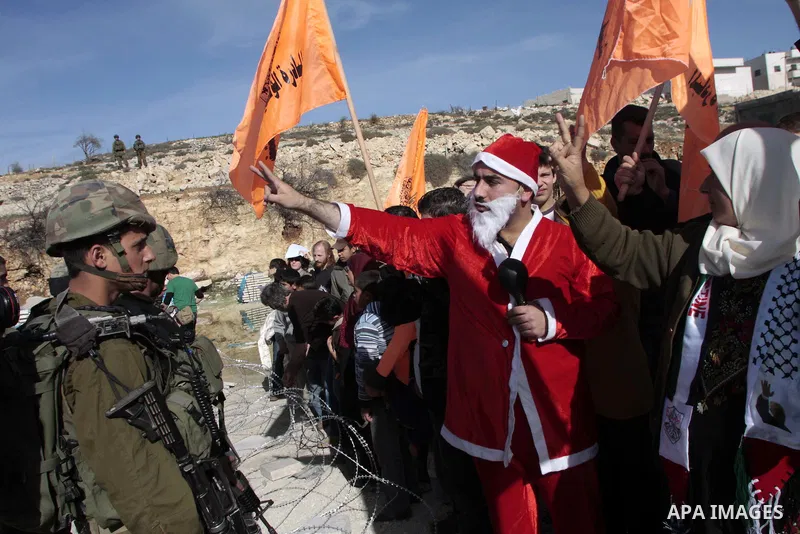 Palestinian protesters dressed in Santa Claus costumes stand opposite Israeli soldiers and behind barbed wires during a demonstration against Israel's controversial separation barrier in the village of Maasarah, near the...