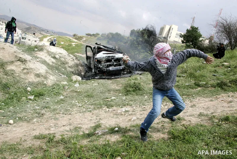 Masked Palestinian youths hurl stones toward Israeli soldiers near a burnt Israeli army vehicle on April 22, 2011 after it flipped over and was set alight by Palestinian youths in the flashpoint neighbourhood of Issawiya...