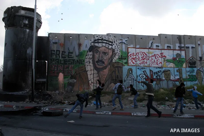 Palestinian youth hurl stones as they stand close to a mural of the late Palestinian leader Yasser Arafat during clashes with Israeli soldiers at the Israeli manned Qalandia checkpoint between Jerusalem and the West Bank...