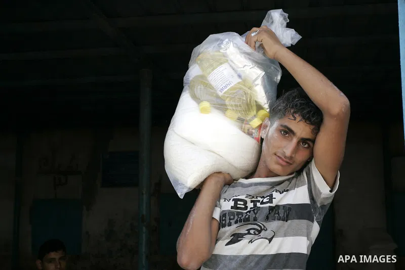 A Palestinian youth carries a bag with wheat flour on his shoulders as others receive food aids from the United Nations Relief and Works Agency (UNRWA) at the Rafah refugee camp, in the southern Gaza Strip on August 15,...