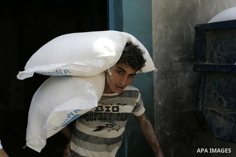 A Palestinian youth carries a bag with wheat flour on his shoulders as others receive food aids from the United Nations Relief and Works Agency (UNRWA) at the Rafah refugee camp, in the southern Gaza Strip on August 15,...