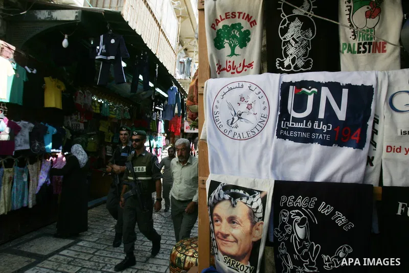 Palestinians and Israeli policemen walk past a souvenir shop in Jerusalem's Old City displaying T-shirts with pictures of French President Nicolas Sarkozy wearing the traditional Arab headdress keffiyeh, as a group of Je...