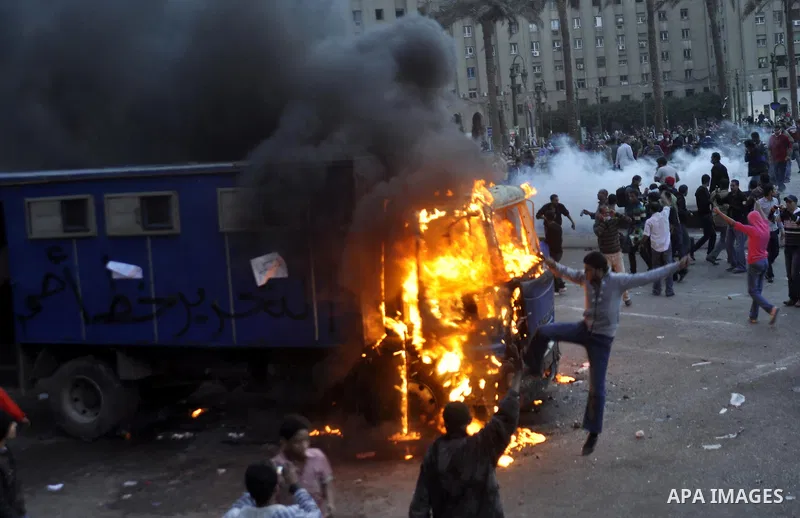 protesters stand next to a police vehicle that was set on fire during clashes between protesters and security forces at Tahrir Square in Cairo November 19, 2011. Clashes erupted between riot police and protesters in Cair...