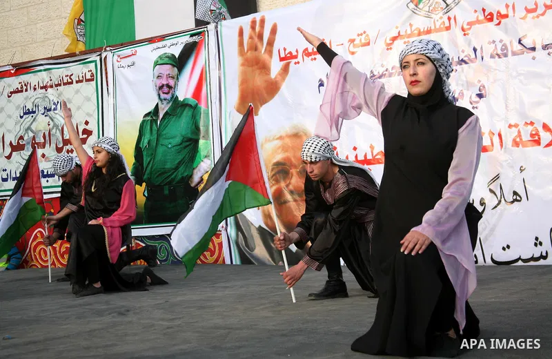 Palestinian folkloric dancers perform in front of a poster showing the late Palestinian leader Yasser Arafat during a celebration to to mark the 47th anniversary of the Fatah political movement. in the West Bank city of...