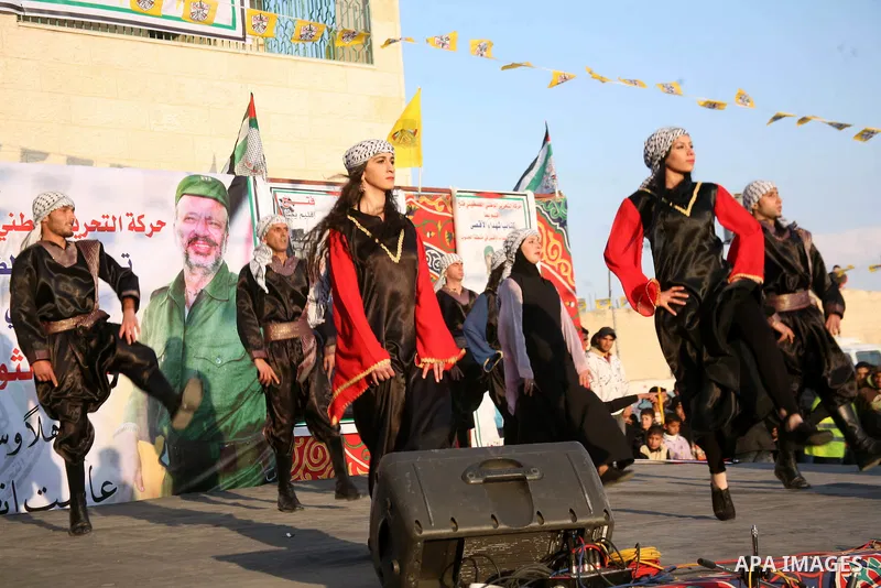 Palestinian folkloric dancers perform in front of a poster showing the late Palestinian leader Yasser Arafat during a celebration to to mark the 47th anniversary of the Fatah political movement. in the West Bank city of...