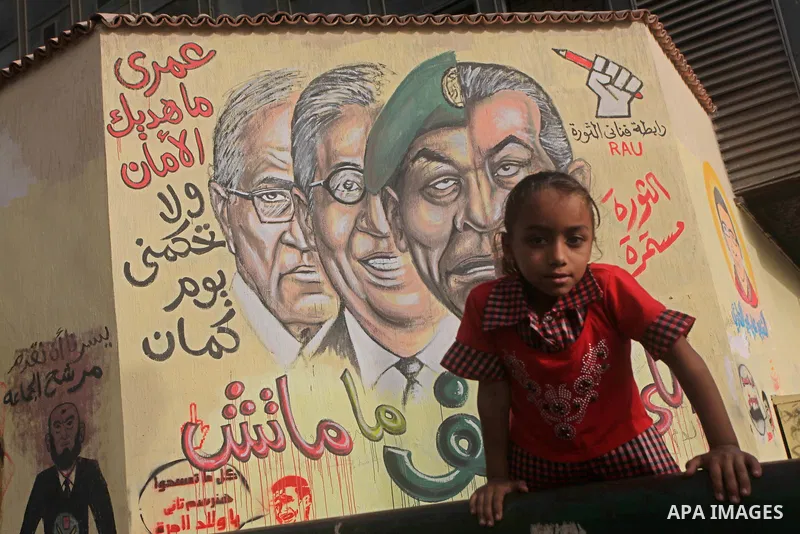 A Egyptian girl sit in front of a mural depicting presidential candidate Ahmed Shafiq (L) and a combination of the faces of former president Hosni Mubarak and Field Marshal Mohamed Hussein Tantawi at Tahrir Square in Cai...