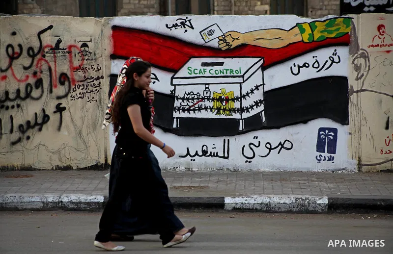 Egyptians walk past graffiti depicting a chained ballot box being controlled by the ruling military council at Tahrir Square in Cairo on May 25, 2012. Egyptians reacted nervously to the first results of their presidentia...