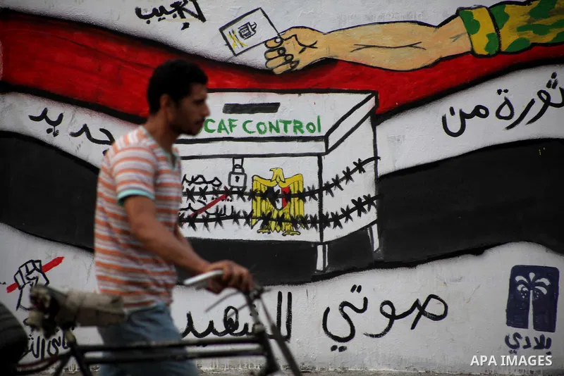 Egyptians walk past graffiti depicting a chained ballot box being controlled by the ruling military council at Tahrir Square in Cairo on May 25, 2012. Egyptians reacted nervously to the first results of their presidentia...