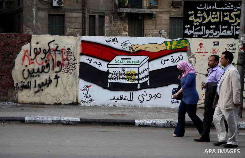 Egyptians walk past graffiti depicting a chained ballot box being controlled by the ruling military council at Tahrir Square in Cairo on May 25, 2012. Egyptians reacted nervously to the first results of their presidentia...
