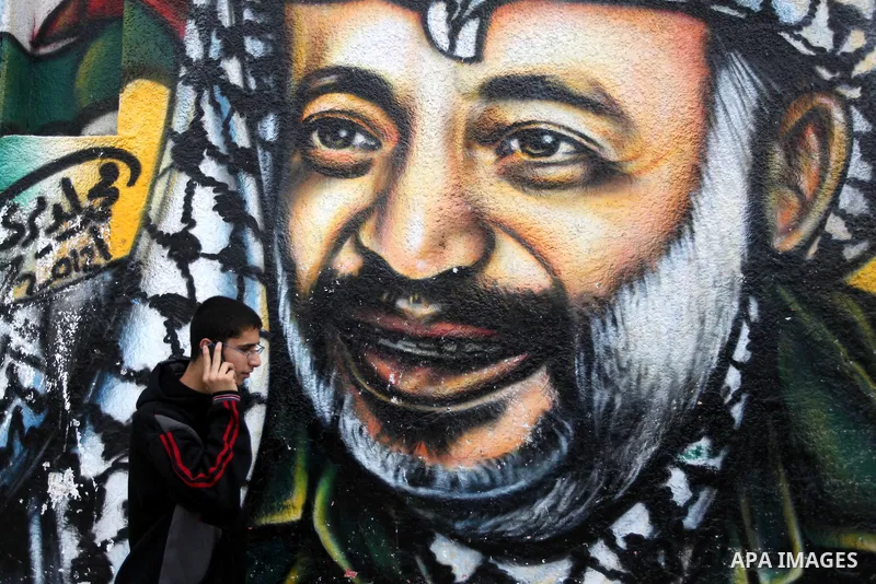 Palestinians walk in front of a mural of late Palestinian leader Yasser Arafat in Gaza City on November 27, 2012. The body of Arafat was exhumed, eight years after his death, as part of an investigation into allegations...