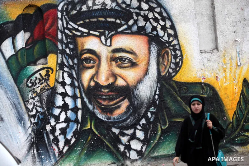 Palestinians walk in front of a mural of late Palestinian leader Yasser Arafat in Gaza City on November 27, 2012. The body of Arafat was exhumed, eight years after his death, as part of an investigation into allegations...