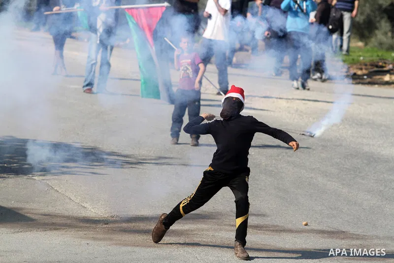 A Palestinian stone-throwing protester wearing a Santa Claus hat, uses a sling to throw back a tear gas canister fired by Israeli security officers (unseen) during clashes at a protest against Jewish settlements, in the...