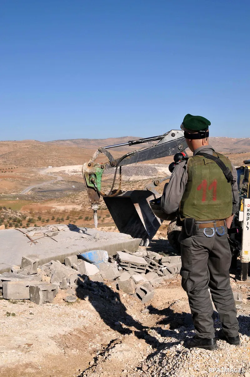 An Israeli solider stands guard as a bulldozer knocks down the wall of a house in the village of Yatta, near the West Bank city of Hebron, 21 January 2013. The house was reportedly located in a closed military zone where...
