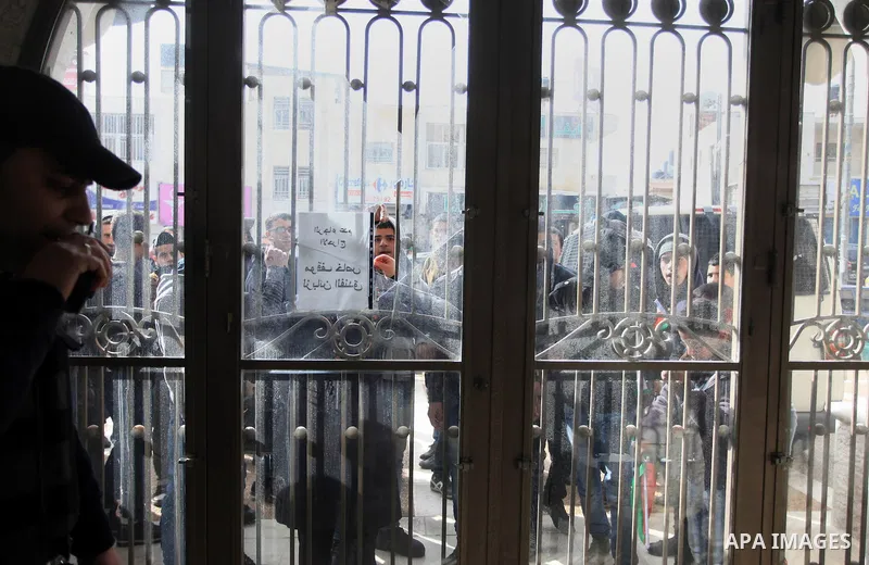 A Palestinian police officer stands guard as Palestinian protesters look through broken glass doors of a gate at a hotel where a group of Israeli and Palestinian peace activists hold a meeting in the West Bank city of Ra...