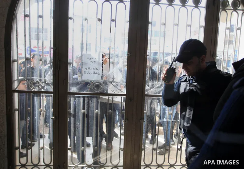 A Palestinian police officer stands guard as Palestinian protesters look through broken glass doors of a gate at a hotel where a group of Israeli and Palestinian peace activists hold a meeting in the West Bank city of Ra...