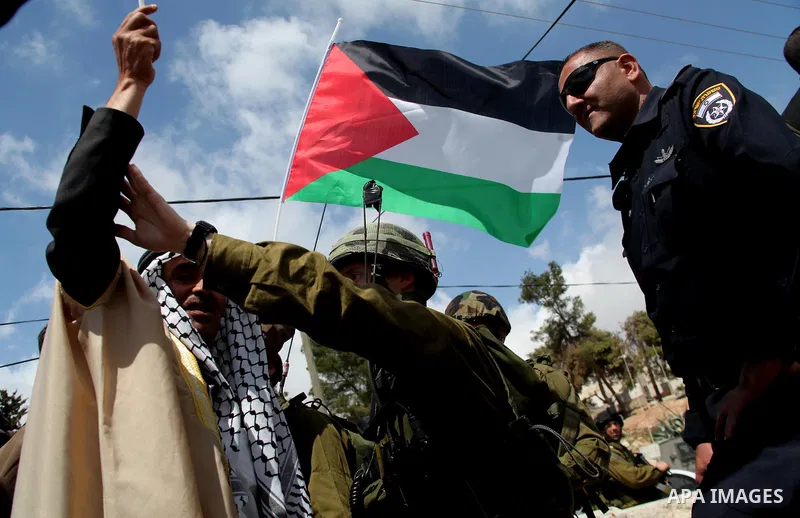 A Palestinian man in traditional garb holds the national flag as he points to an Israeli soldiers during a protest near an Israeli-occupied Palestinian home, located close to the Kiryat Arbaa Jewish settlment, as Palesti...