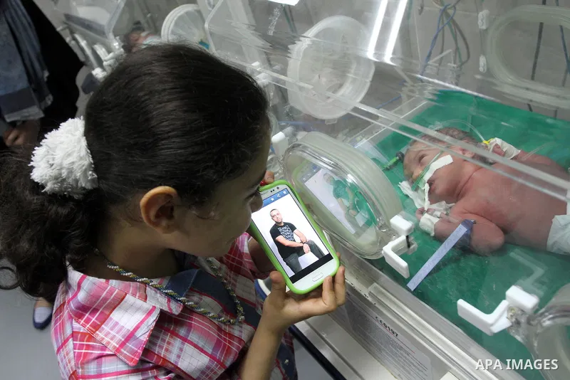 Aseel Khadoora, the daughter of Palestinian prisoner Saleh Khadoora (pictured in mobile), stands near her new brother as he lies in an incubator at a hospital, in Gaza City on June 26, 2014. The wife of Palestinian jaile...