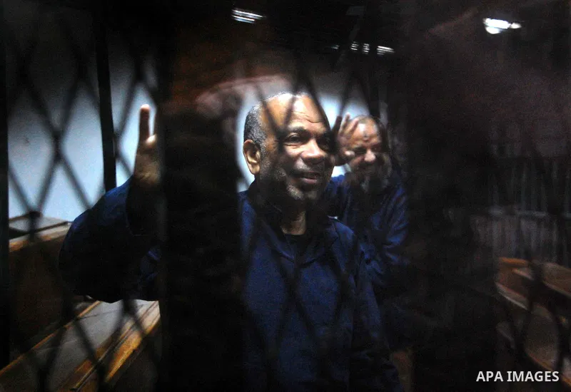 Egyptian Muslim Brotherhood top figures, Saad el-Katatni and Mohamed el-Beltagy flash the four finger symbol known as "Rabaa" from behind the defendant's cage during his trial for insulting the judiciary alongside 25 oth...