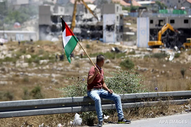 A Palestinian man holds a Palestinian flag as Israeli heavy machinery demolish vacant apartment blocs by order of Israel's high court, in the West Bank Jewish settlement of Beit El near Ramallah July 29, 2015. Israel gav...