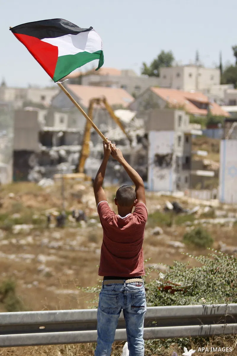 A Palestinian man holds a Palestinian flag as Israeli heavy machinery demolish vacant apartment blocs by order of Israel's high court, in the West Bank Jewish settlement of Beit El near Ramallah July 29, 2015. Israel gav...