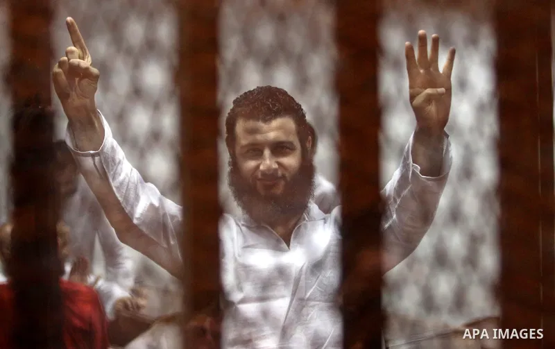 An Egyptian Muslim Brotherhood defendant waves as he stands behind the bars during his trial, along with ousted Islamist president Mohamed Morsi (unseen), in a court in Cairo on August 8, 2015. Cairo Criminal Court resum...