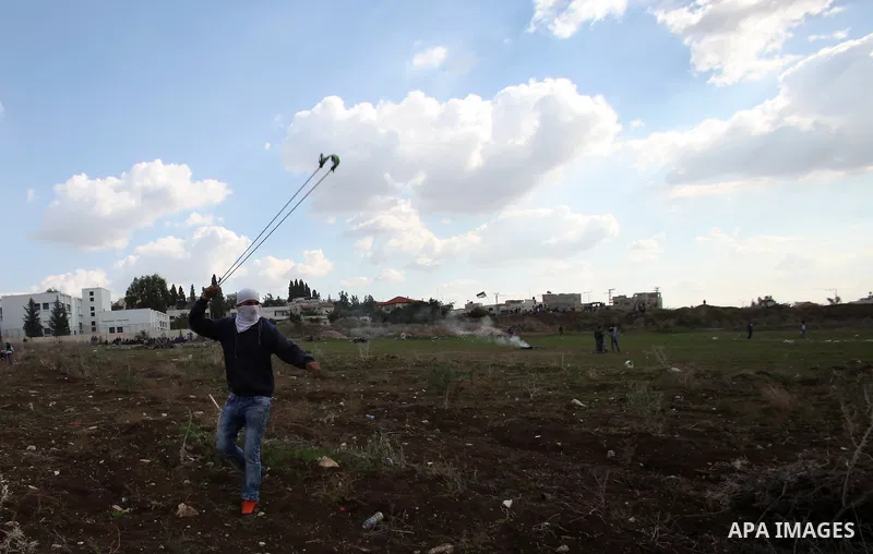 A Palestinian protester uses a sling shot to throw stones towards Israeli security forces during clashes following a protest to mark the anniversary of the death of late Palestinian leader Yasser Arafat, in the West Bank...