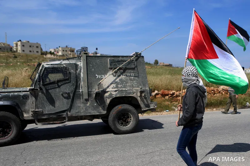 A Palestinian girl waves national flags past Israeli security forces jeep during clashes following a march against Palestinian land confiscation on April 15, 2016 in the West Bank village of Nabi Saleh near Ramallah. Pho...
