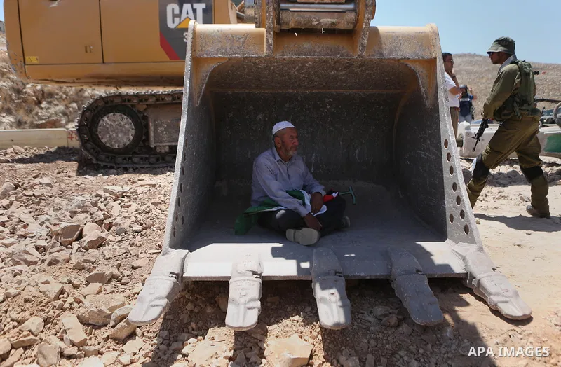 A man holding a Palestinian flag protests as he sits in the scoop of an Israeli excavator as tries to prevent it from clearing his land during a protest against Jewish settlements, near the village of Deir Qaddis near th...