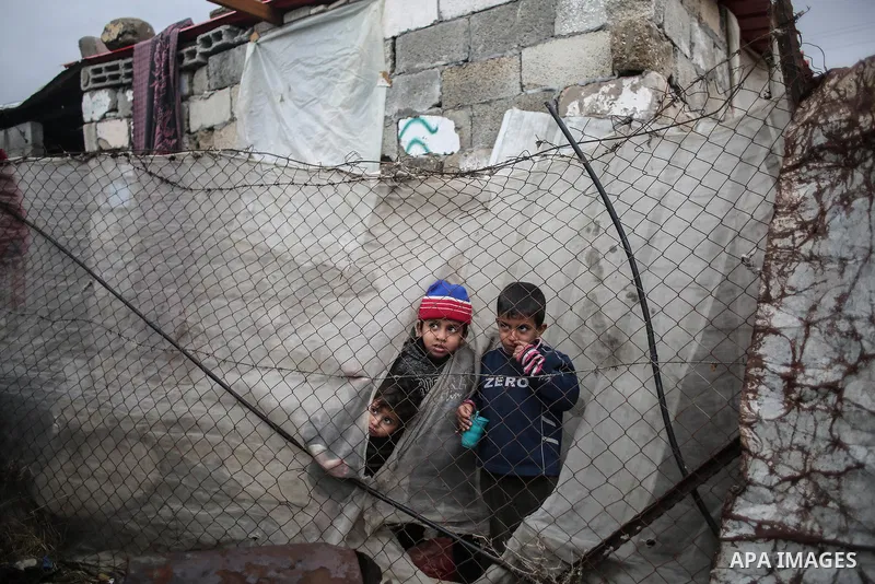 Palestinian children stand between a fence and a sheet of cloth surrounding their family house during rainy, cold weather in a slum on the outskirts of Khan Younis refugee camp, southern Gaza Strip, on December 28, 2016....