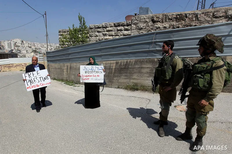 Palestinian demonstrators hold placards near Israeli forces during a protest against Jewish settlements in the West Bank city of Hebron March 26, 2017. Photo by Wisam Hashlamoun