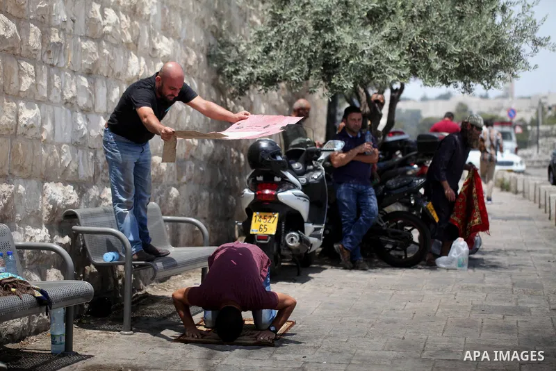 A Palestinian man uses a piece of cardboard to cover a man from the soaring heat as he pray outside the Lion's Gate a main entrance to the Al-Aqsa mosque compound in Jerusalem's Old City on July 24, 2017 as Palestinians...