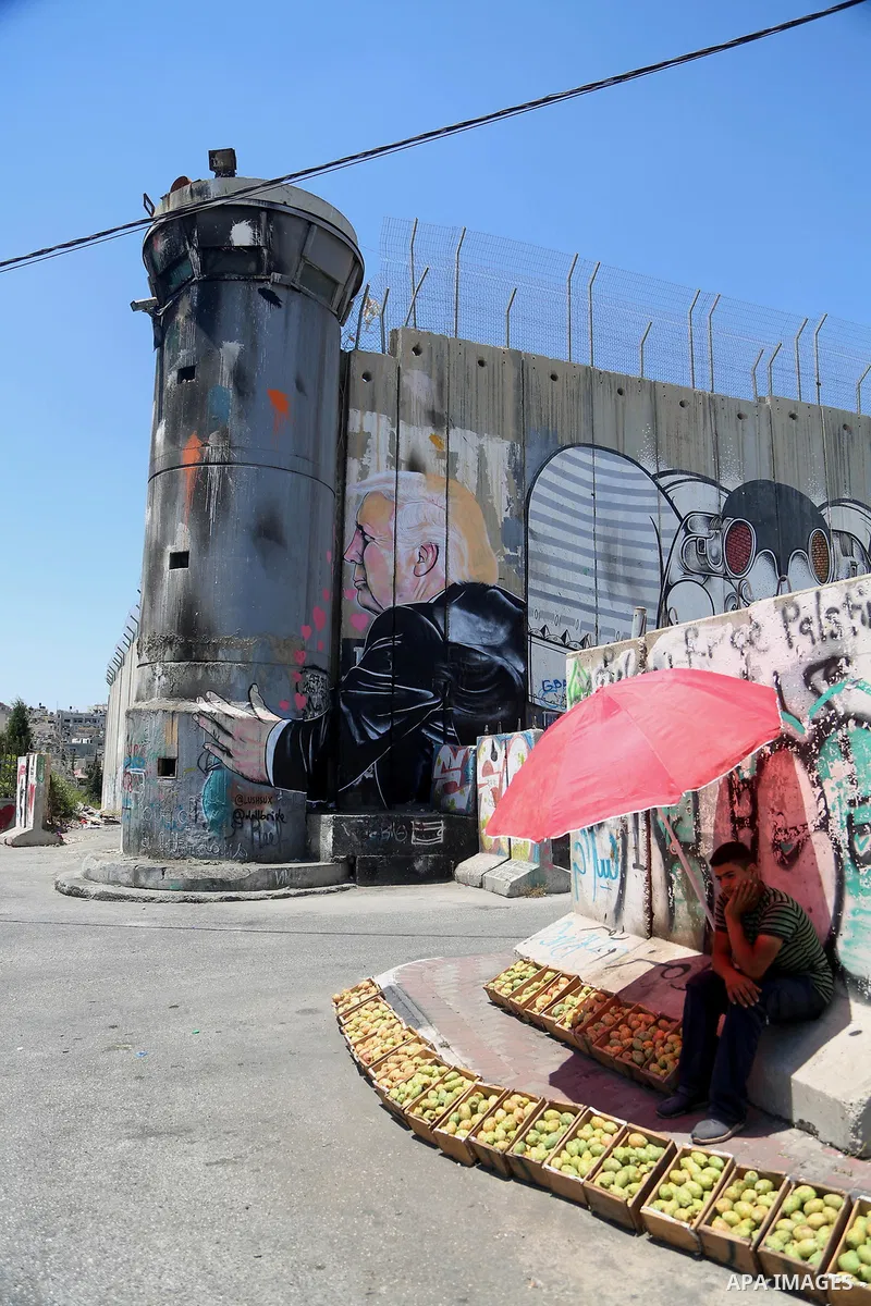 A Palestinian vendor sells vegetables in front of a graffiti depicting US President Donald Trump on the controversial Israeli separation barrier in the West Bank town of Bethlehem on August 8, 2017. Photo by Shadi Hatem