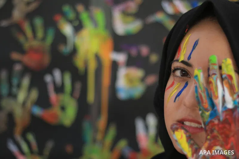A young girl with hijab uses a colored hand to cover one eye. Photo by Sanad Ltefa