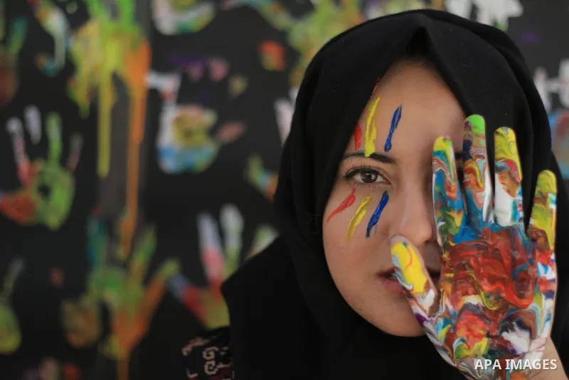 A young girl with hijab uses a colored hand to cover one eye. Photo by Sanad Ltefa