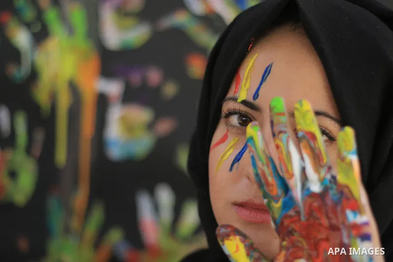 A young girl with hijab uses a colored hand to cover one eye. Photo by Sanad Ltefa