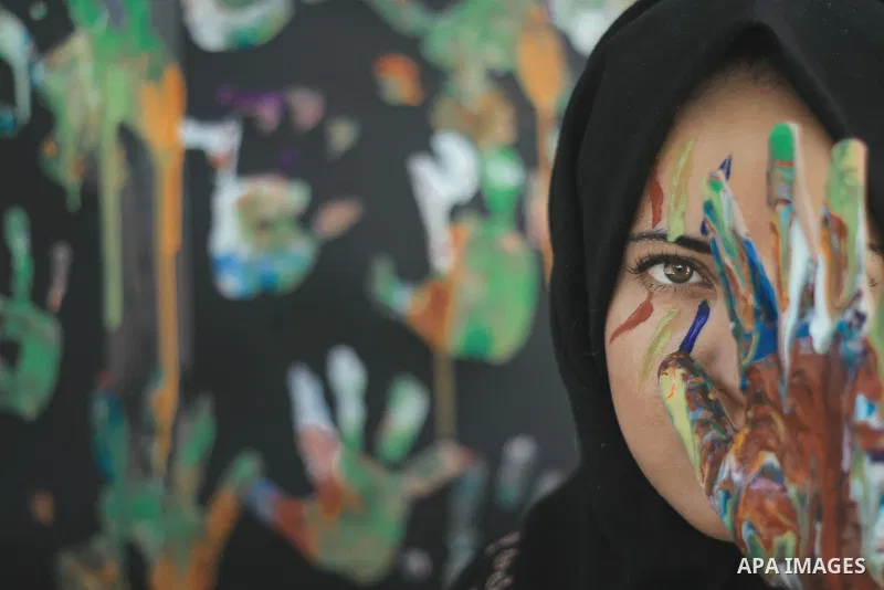 A young girl with hijab uses a colored hand to cover one eye. Photo by Sanad Ltefa