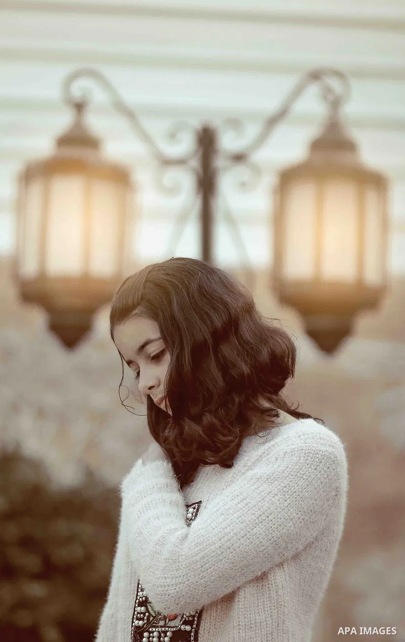 A pretty girl puts one hand at her shoulder with lamps at background. Photo by Sanad Ltefa