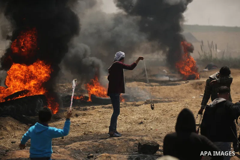 A Palestinian protester uses a slingshot to hurl stones as others burn tires to protect themselves from shots of Israeli soldiers during clashes at the Israel-Gaza border in a tent city protest demanding the right to ret...