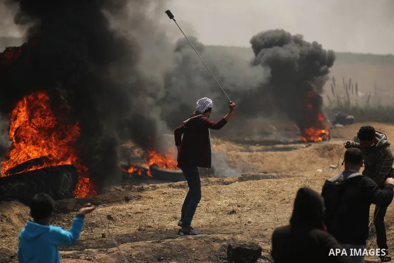 A Palestinian protester uses a slingshot to hurl stones as others burn tires to protect themselves from shots of Israeli soldiers during clashes at the Israel-Gaza border in a tent city protest demanding the right to ret...