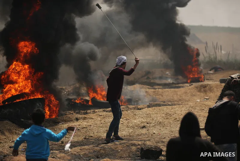 A Palestinian protester uses a slingshot to hurl stones as others burn tires to protect themselves from shots of Israeli soldiers during clashes at the Israel-Gaza border in a tent city protest demanding the right to ret...