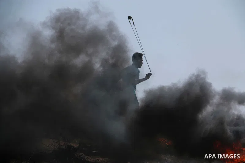 A Palestinian protester uses a slingshot to hurl stones as other burn tires to protect themselves from shots of Israeli soldiers during clashes in a tent city protest at the Israel-Gaza border, demanding the right to ret...