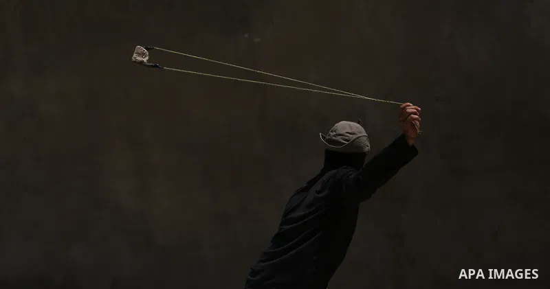 A masked Palestinian uses a slingshot to hurl stones towards Israeli troops during clashes following a demonstration against the expropriation of Palestinian land by Israel in the village of Kfar Qaddum, near Nablus, in...