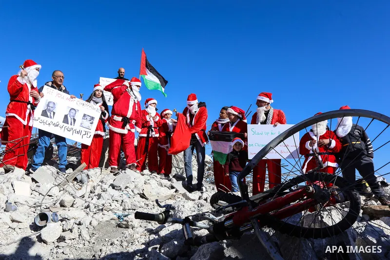 A Palestinian dressed as Santa Claus hands out gifts to children sitting on the remains of a demolished Palestinian house during an anti-Israel protest, in the West Bank city of Bethlehem, on December 23, 2019. Photo by...