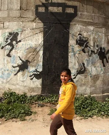 A Palestinian girl walks next to a mural of children using an Israeli army watch tower as a swing ride, said to have been painted by British street artist Banksy, on the remains of a house that was destroyed during the 5...