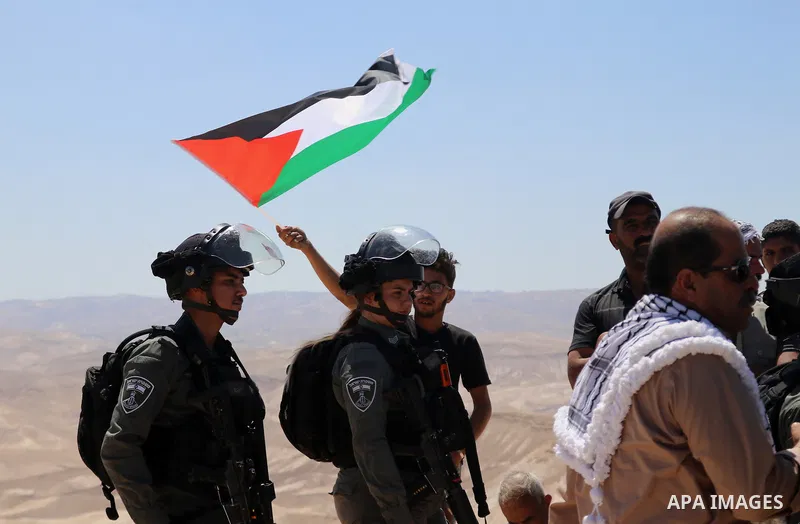 Palestinian demonstrators hold placards and national flags in front of Israeli soldiers during a protest against Israeli settlements near Hebron in the Israeli-occupied West Bank June 4, 2021. Photo by Mosab Shawer