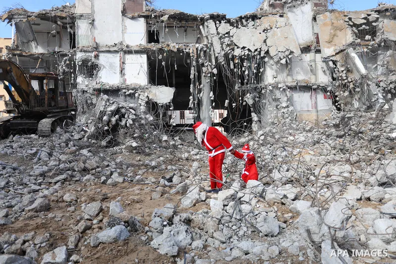 A Palestinian man and his son dressed as Santa Claus walk near the rubbles of al-Jawhara tower which was hit by Israeli strikes during the recent 11-day conflict between Israel and Hamas on May, on Christmas Eve, in Gaza...
