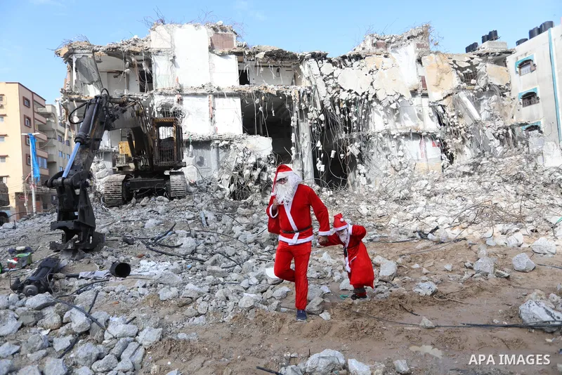 A Palestinian man and his son dressed as Santa Claus walk near the rubbles of al-Jawhara tower which was hit by Israeli strikes during the recent 11-day conflict between Israel and Hamas on May, on Christmas Eve, in Gaza...