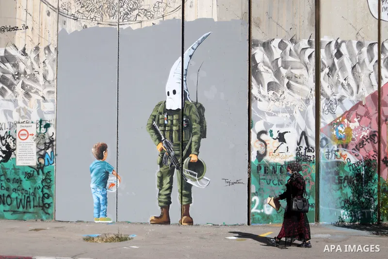 A Palestinian woman walks past a graffiti mural depicting a boy holding a fishbowl looking at a uniformed Israeli soldier wearing a white hood in the style of the Ku Klux Klan (KKK), drawn by Palestinian artist Taqi Spat...