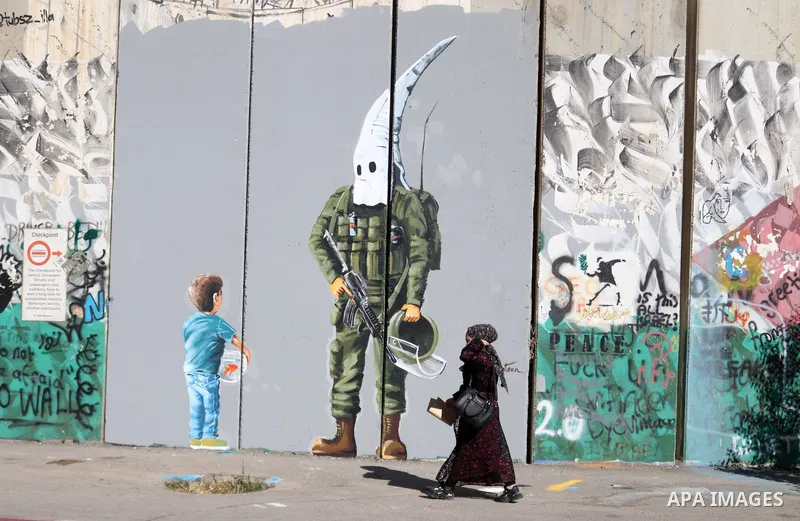 A Palestinian woman walks past a graffiti mural depicting a boy holding a fishbowl looking at a uniformed Israeli soldier wearing a white hood in the style of the Ku Klux Klan (KKK), drawn by Palestinian artist Taqi Spat...