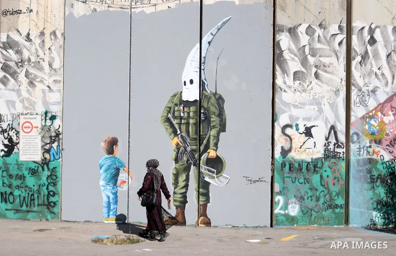A Palestinian woman walks past a graffiti mural depicting a boy holding a fishbowl looking at a uniformed Israeli soldier wearing a white hood in the style of the Ku Klux Klan (KKK), drawn by Palestinian artist Taqi Spat...